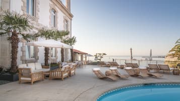 A poolside area with lounge chairs and umbrellas, a building with stone facade, and a view of the sea.