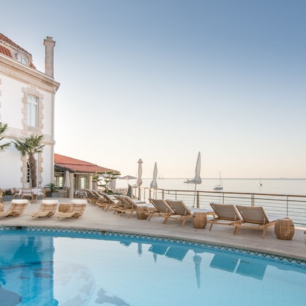 A poolside area with lounge chairs and umbrellas, a building with a red-tiled roof, and a view of the sea.