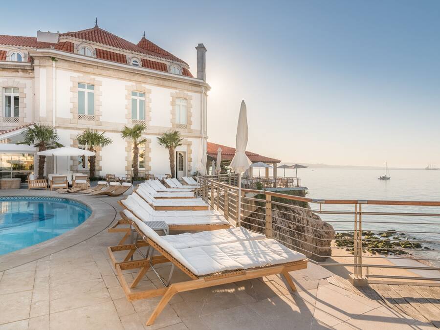 Ein Poolbereich mit Liegestühlen und Sonnenschirmen, ein Gebäude mit einem Ziegeldach und Blick auf das Meer.