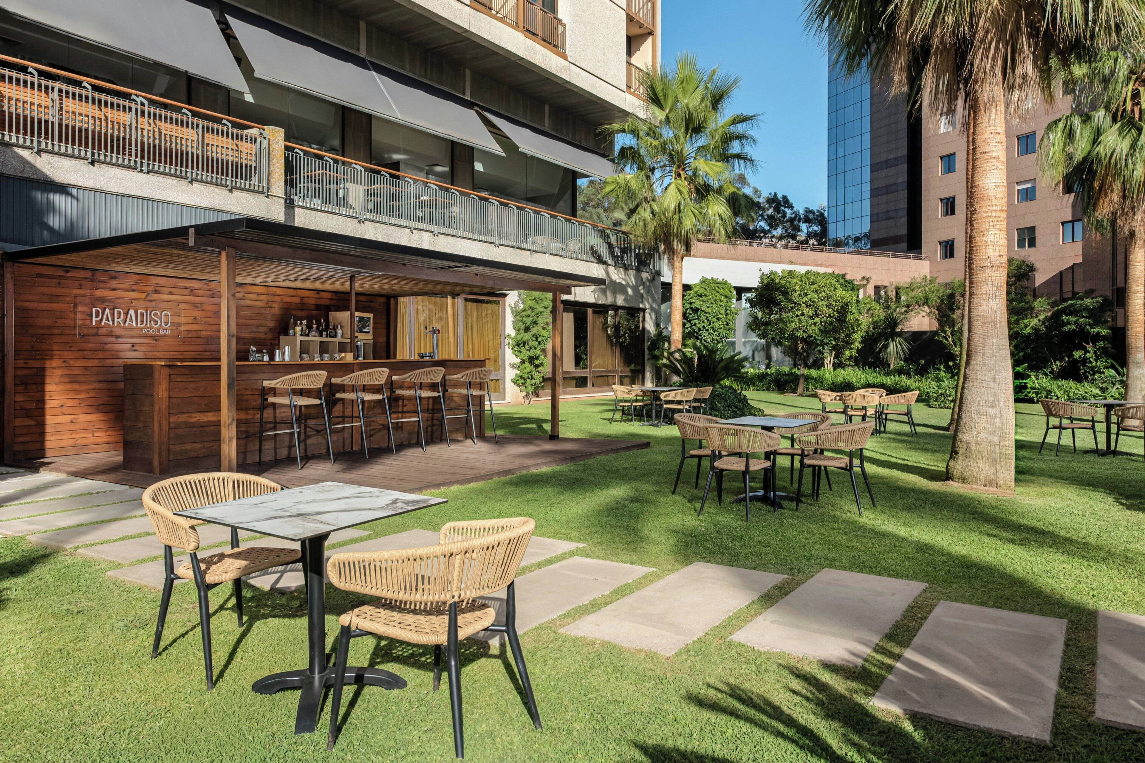 Um pátio com área de estar externa, um bar chamado 'PARADISO' e um edifício moderno ao fundo.
