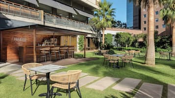 Um pátio com área de estar externa, um bar chamado 'PARADISO' e um edifício moderno ao fundo.