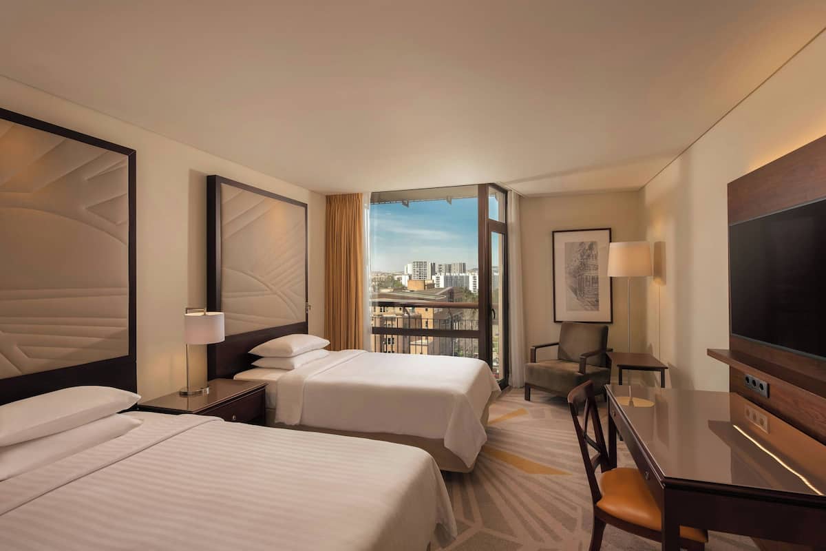 A hotel room with two beds, a TV, a desk, and a view of the city.