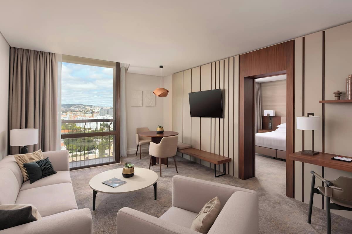 A modern hotel room with a large window, a flat-screen TV, a sofa, a coffee table, a desk, and a bed.