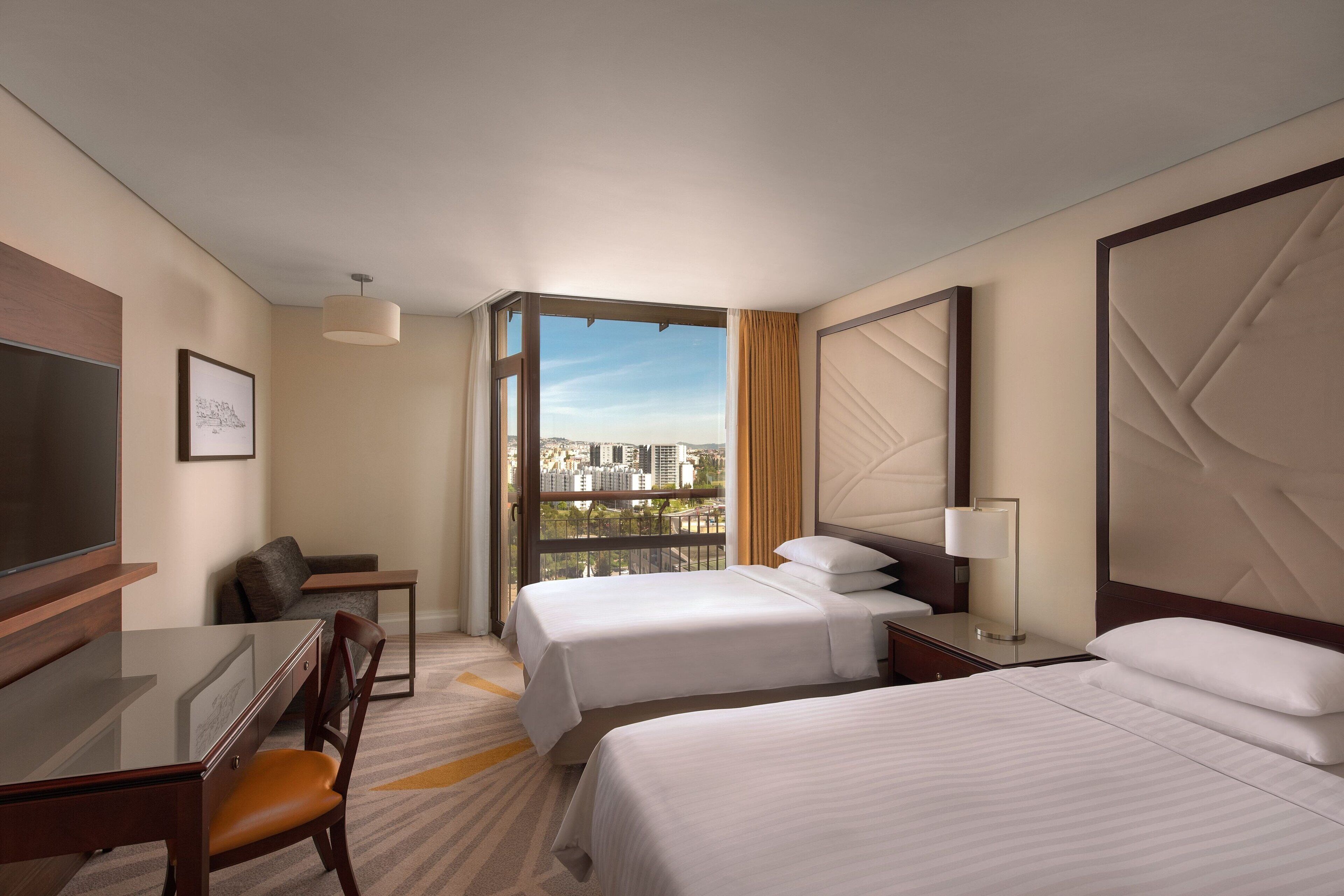 A hotel room with two beds, a desk, a chair, a TV, and a large window with a city view.