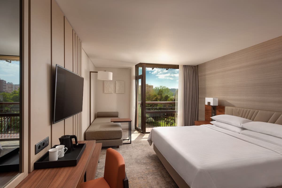 A modern hotel room with a large bed, a desk with a coffee maker, a flat-screen TV, and a balcony with a city view.