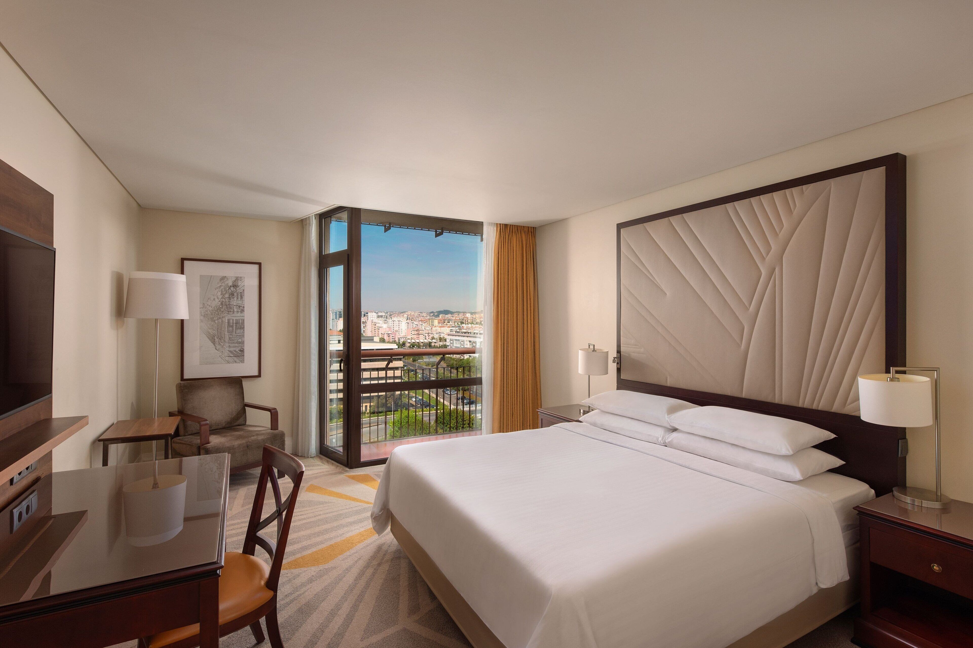 A hotel room with a bed, a desk, a chair, a TV, and a balcony with a city view.