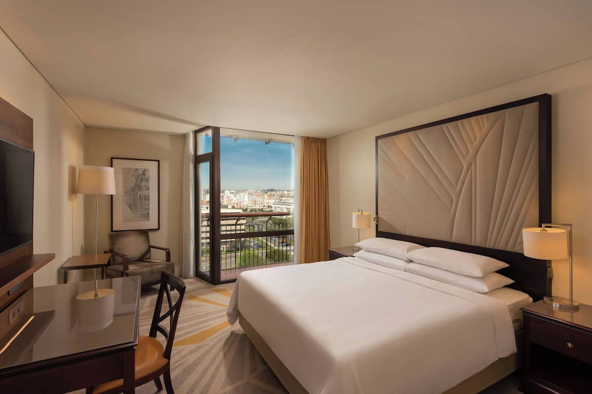 A hotel room with a bed, a desk, a chair, a TV, and a large window offering a city view.