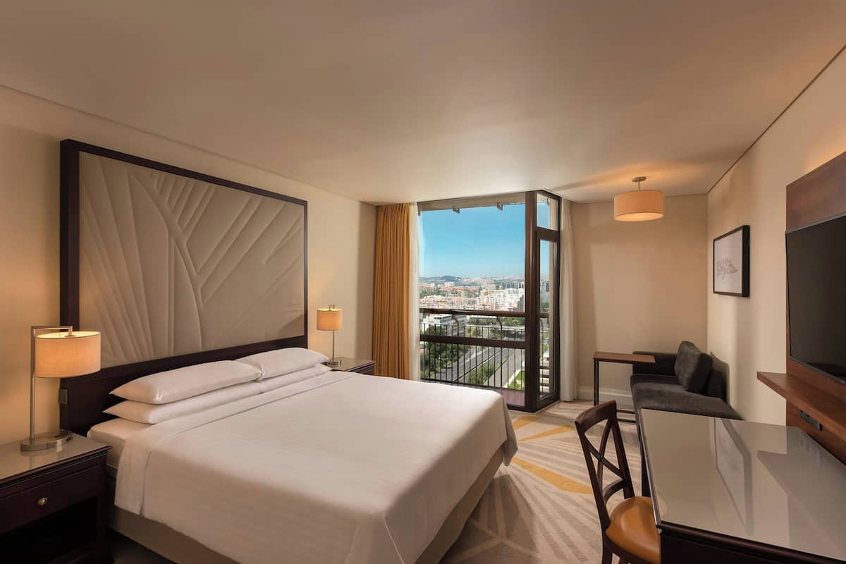 A hotel room with a large bed, a desk, a chair, a TV, and a balcony with a city view.