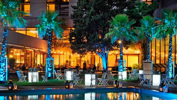 Un área junto a la piscina con sillones y mesas, rodeada de palmeras y un edificio con ventanas iluminadas.