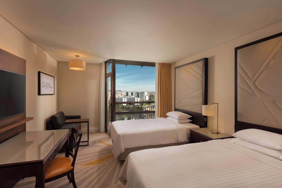 A hotel room with two beds, a desk, a chair, a TV, and a large window with a city view.