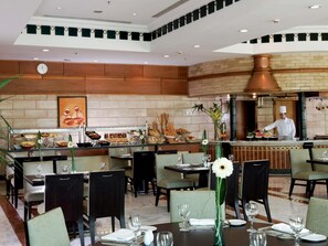 A restaurant with a buffet, dining tables, chairs, and a chef preparing food. - Mövenpick Cairo Media City (6th of October City)