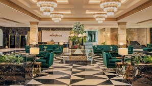 A lobby with a geometric patterned floor, green upholstered sofas, chandeliers, a reception desk, and a large floral arrangement. - Mövenpick Cairo Media City (6th of October City)