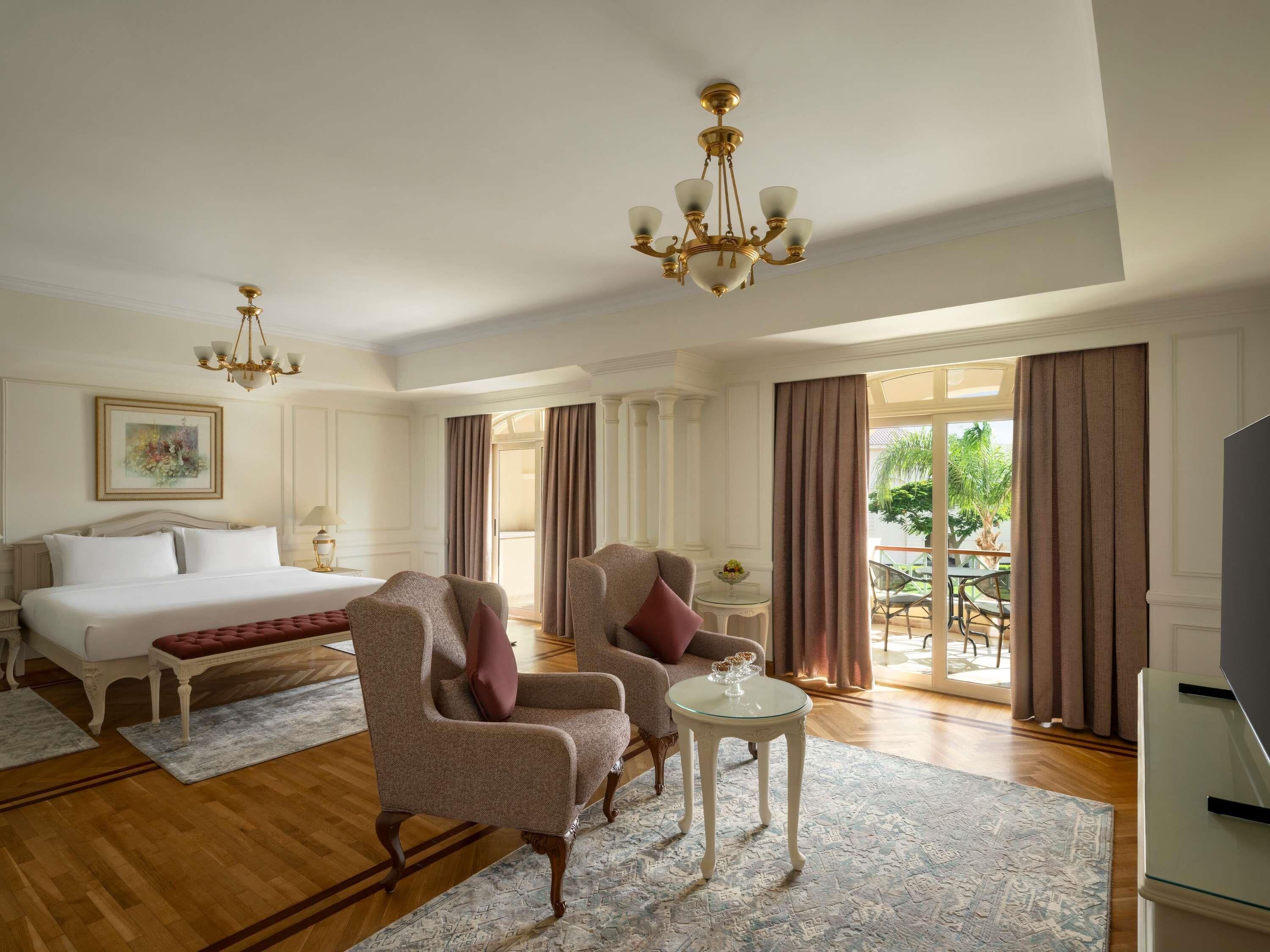 A hotel room with a bed, two armchairs, a small side table, a television, and a large window with curtains.