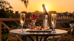 A table with a bottle of champagne, two glasses of champagne, a bowl of strawberries, and a plate of appetizers on a balcony overlooking a sunset. - Mövenpick Cairo Media City (6th of October City)
