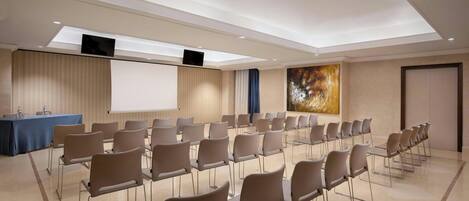 Uma sala de conferências com fileiras de cadeiras, uma tela de projeção e um grande quadro abstrato na parede.
