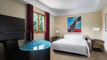 A hotel room with a bed, a desk, a chair, a window with curtains, a painting on the wall, and a lamp on the nightstand.
