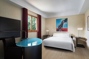 A hotel room with a bed, a desk, a chair, a window with curtains, a painting on the wall, and a lamp on the nightstand.