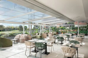 Un área de comedor al aire libre amplia con paredes de vidrio, asientos acolchados y mesas dispuestas para los huéspedes.