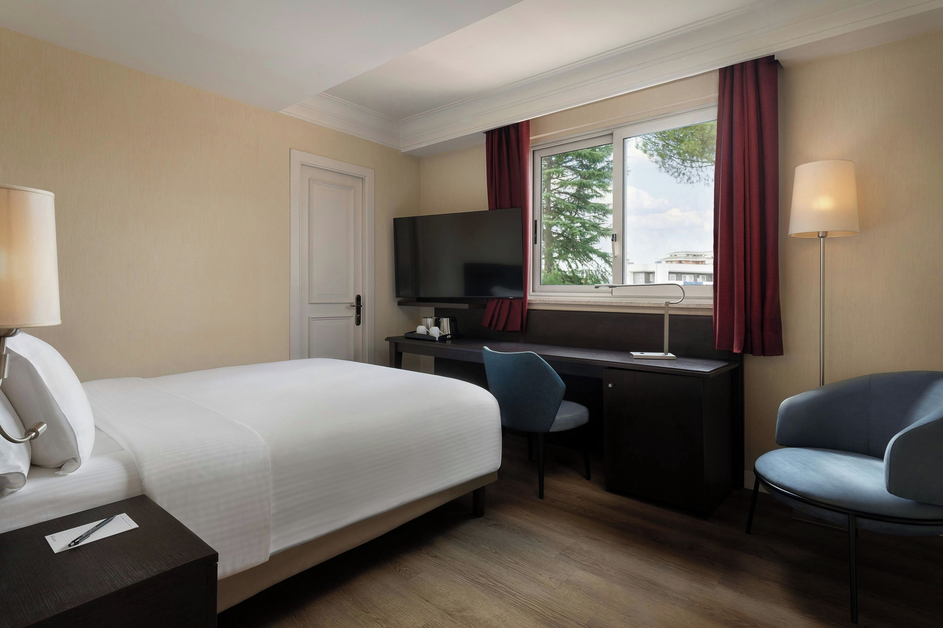 Habitación de hotel con cama, escritorio, silla, televisión y ventana con cortinas.