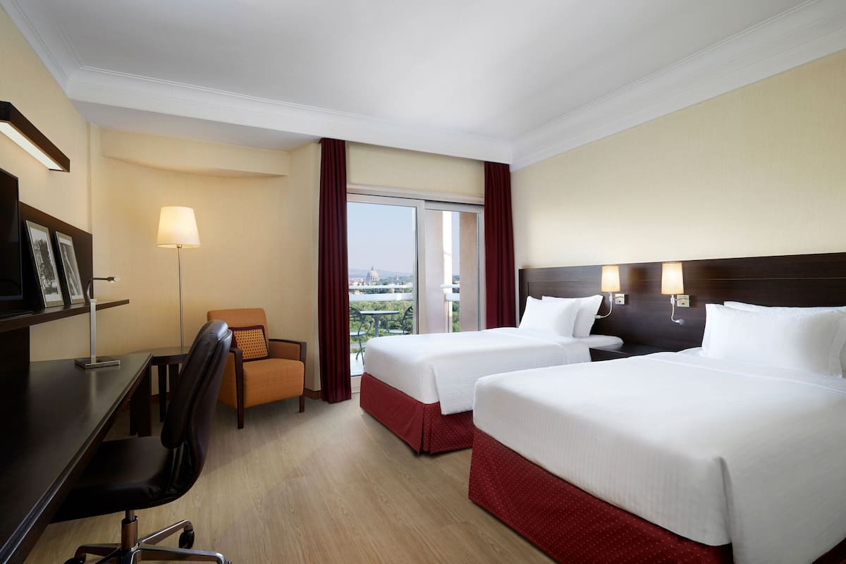 A hotel room with two beds, a desk, a chair, a lamp, and a view of the cityscape.