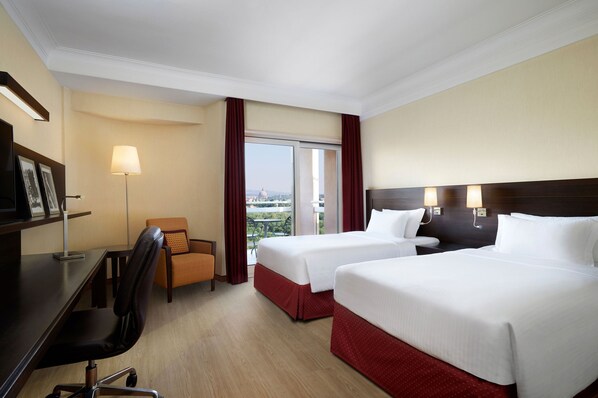 A hotel room with two beds, a desk, a chair, a lamp, and a view of the cityscape. - Courtyard by Marriott Rome Central Park (Rome)