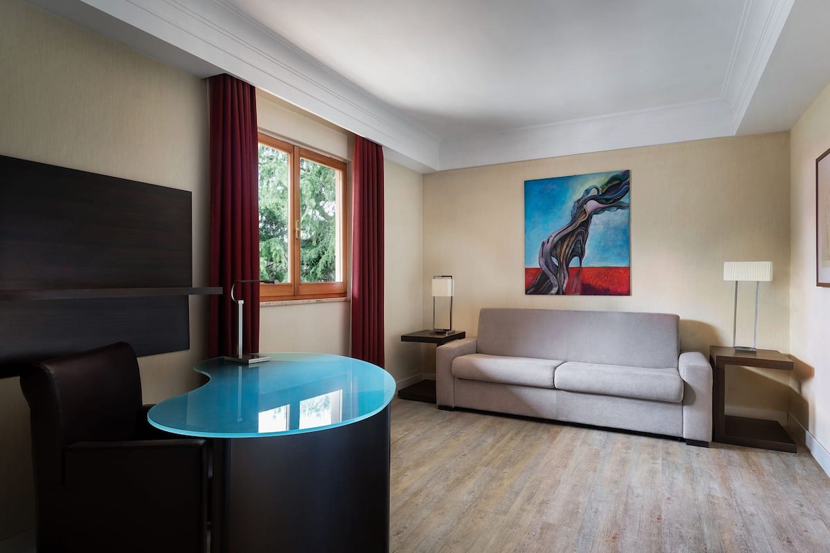 Ein modernes Hotelzimmer mit Glastisch, einer Couch, einem Bild an der Wand und einem Fenster mit Vorhängen.