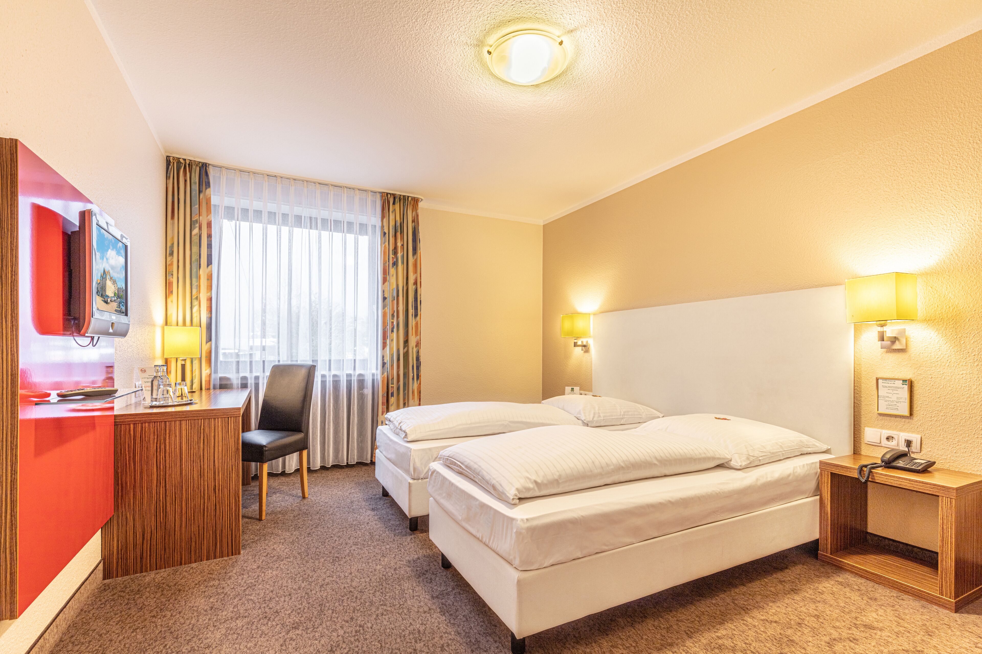 Habitación de hotel con dos camas, un escritorio con silla, televisión y ventana con cortinas.