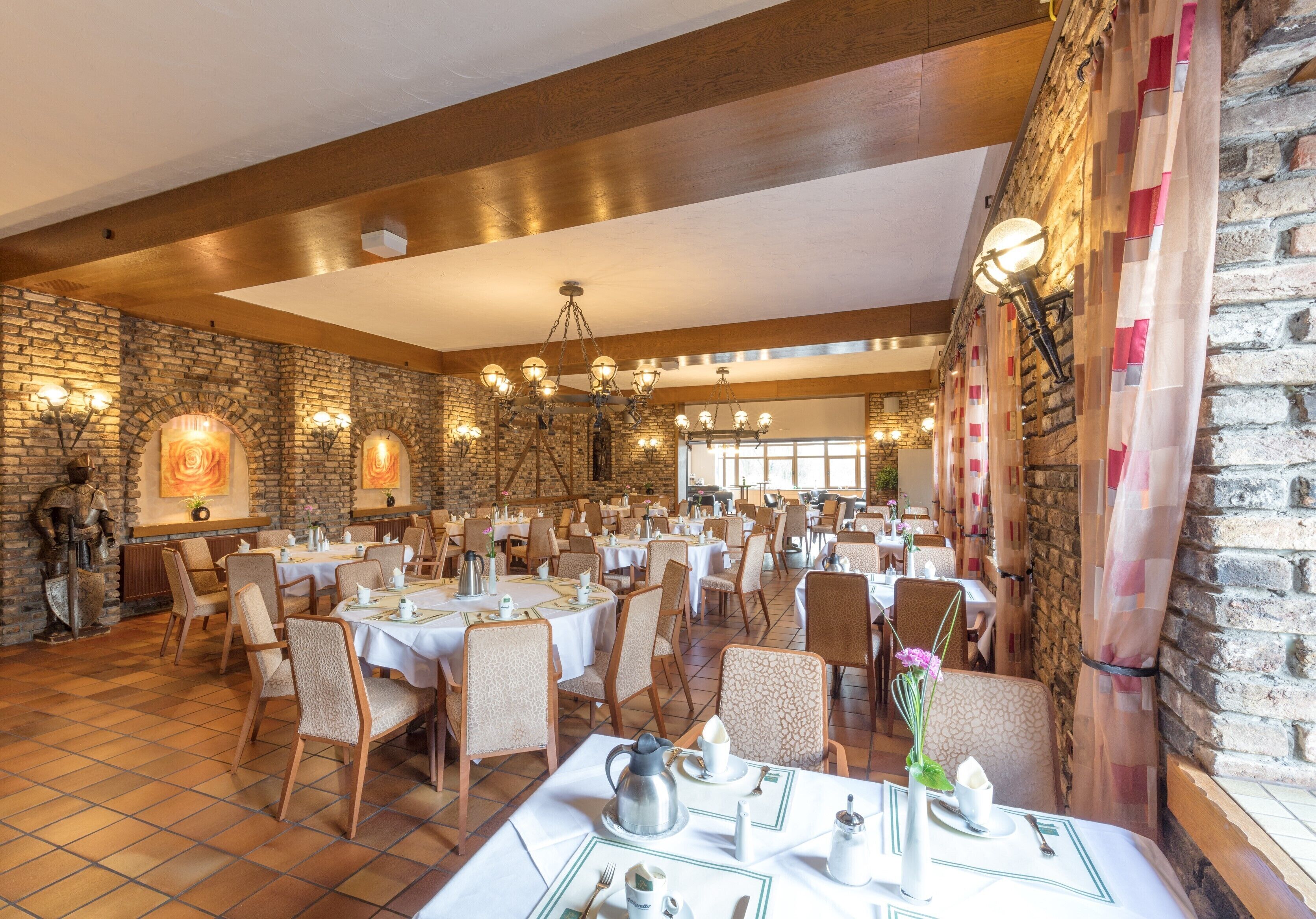 Une vaste salle à manger aux murs de briques, avec des poutres en bois et de nombreuses tables dressées pour le repas.