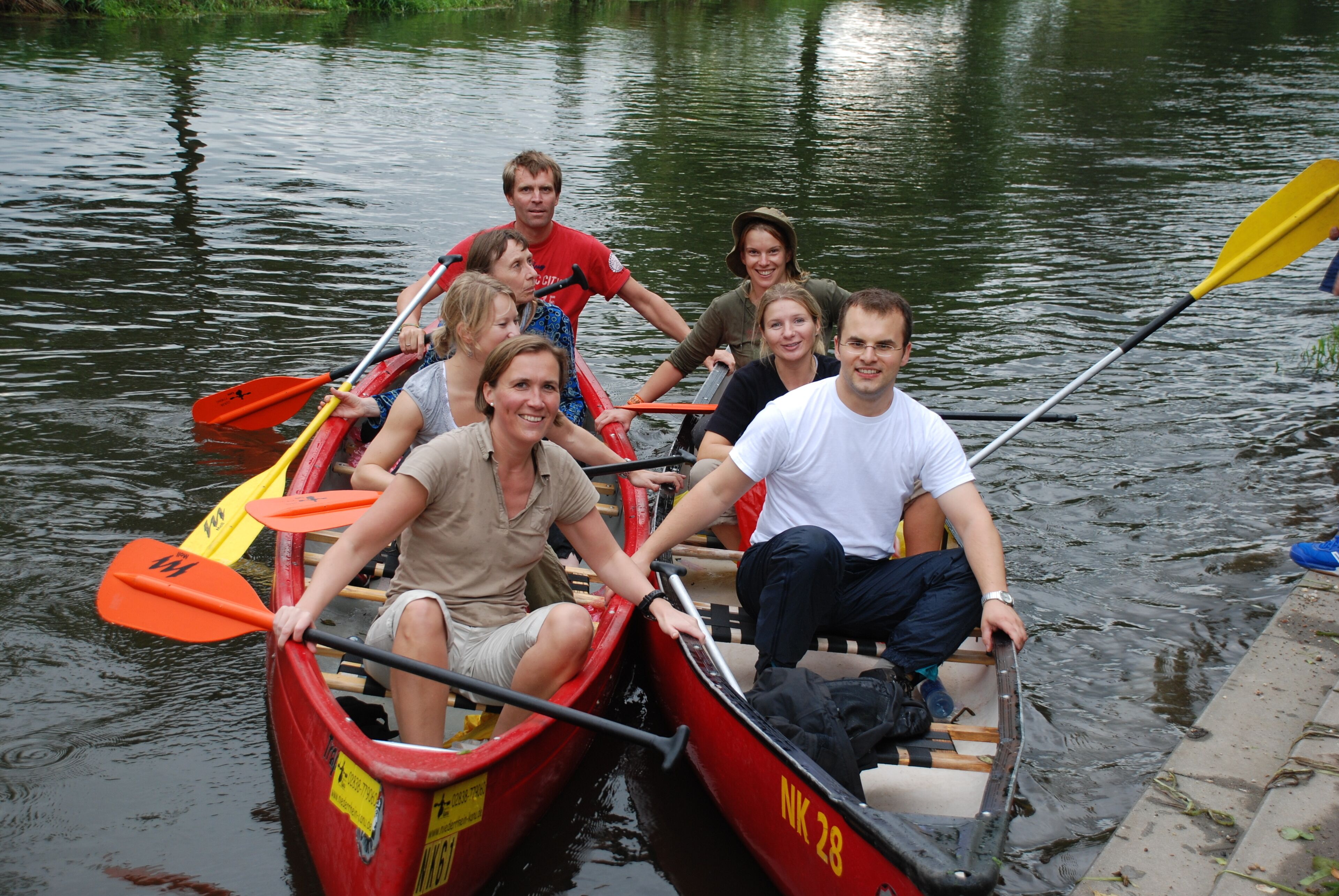 Un groupe de personnes se trouve dans un petit bateau rouge, équipé de rames jaunes, et navigue sur une rivière paisible.