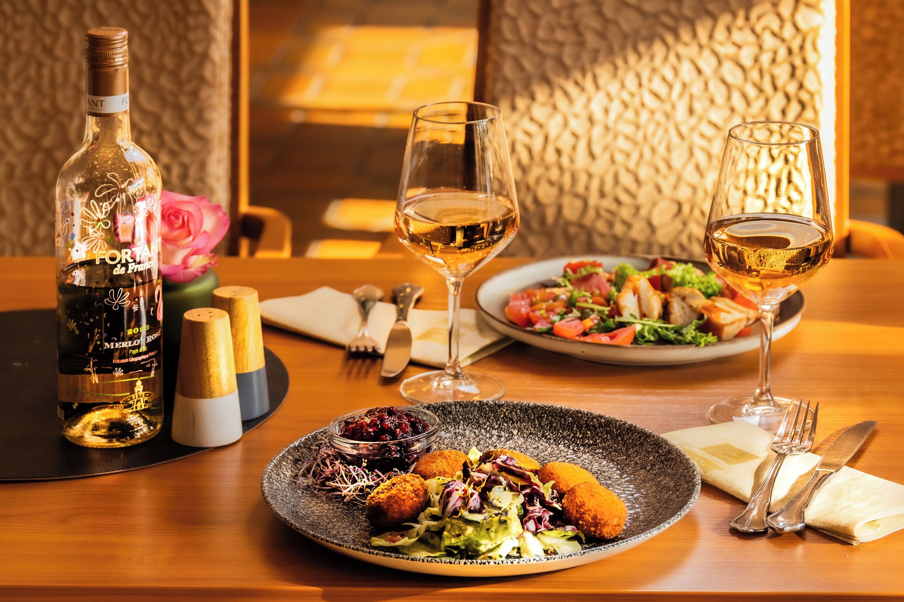 A dining table with a bottle of wine, two glasses of wine, a plate of food, and a rose.