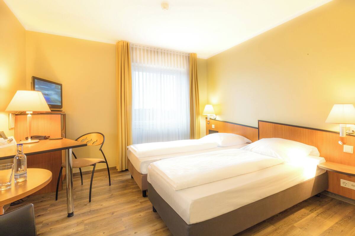 A hotel room with two beds, a desk with a lamp, a chair, and a window with curtains.
