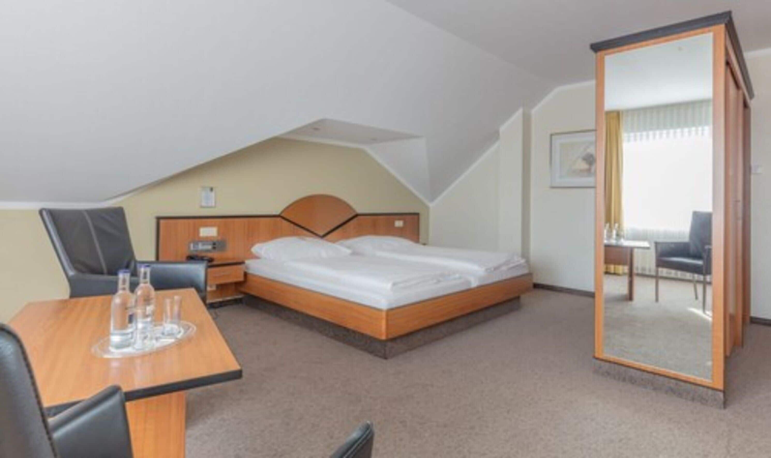 Quarto de hotel com uma cama de solteiro, uma escrivaninha com cadeira, uma mesinha com dois copos e um espelho.