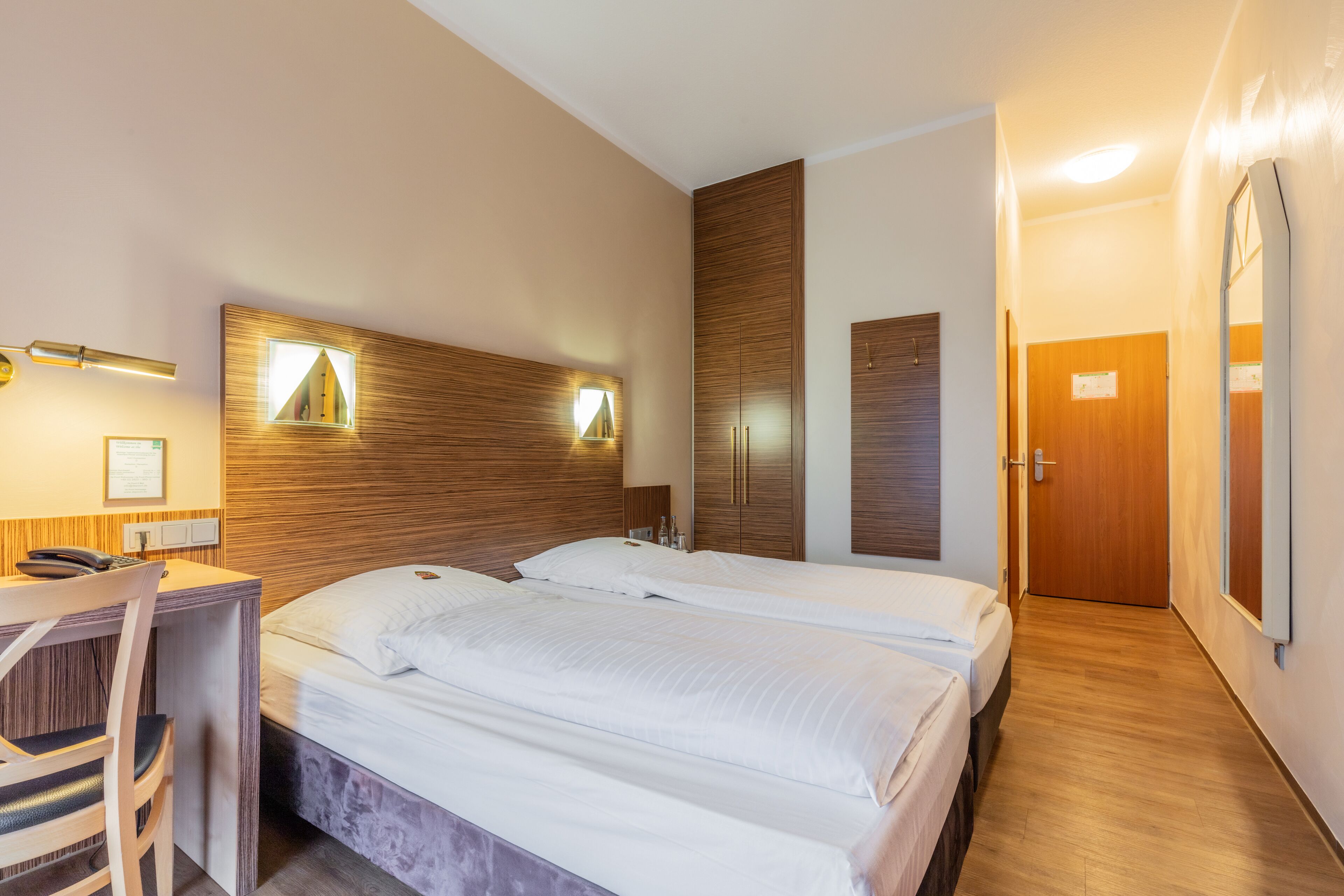 Quarto de hotel com cabeceira de madeira, duas camas, uma escrivaninha com cadeira e uma porta.