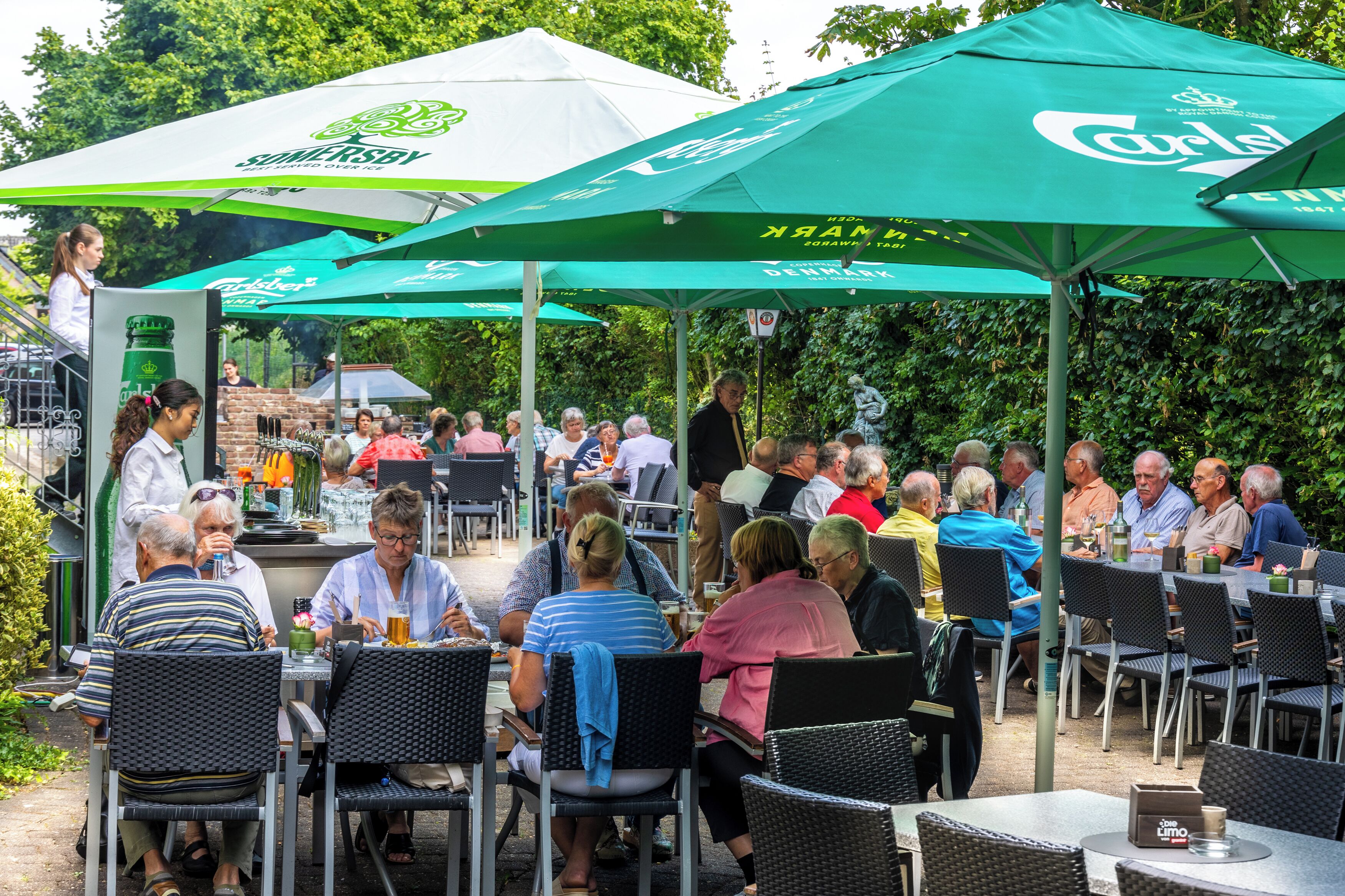 An outdoor dining area with tables and chairs under green umbrellas, a grill in the background, and a statue of a thinker.