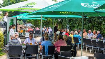An outdoor dining area with tables and chairs under green umbrellas, a grill in the background, and a statue of a thinker.
