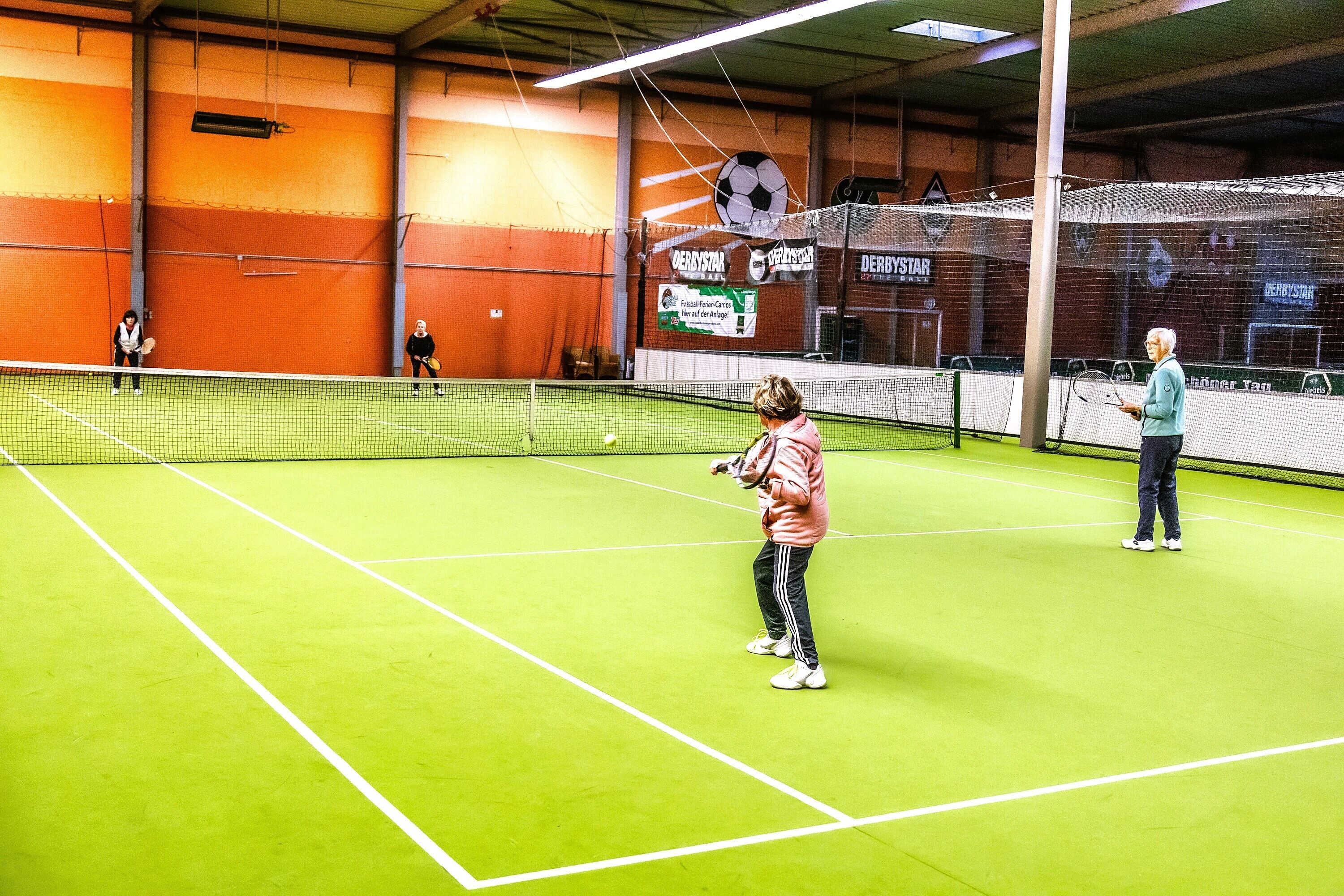 Een indoor tennisbaan met een net, een scorebord en een muur met vlaggen.