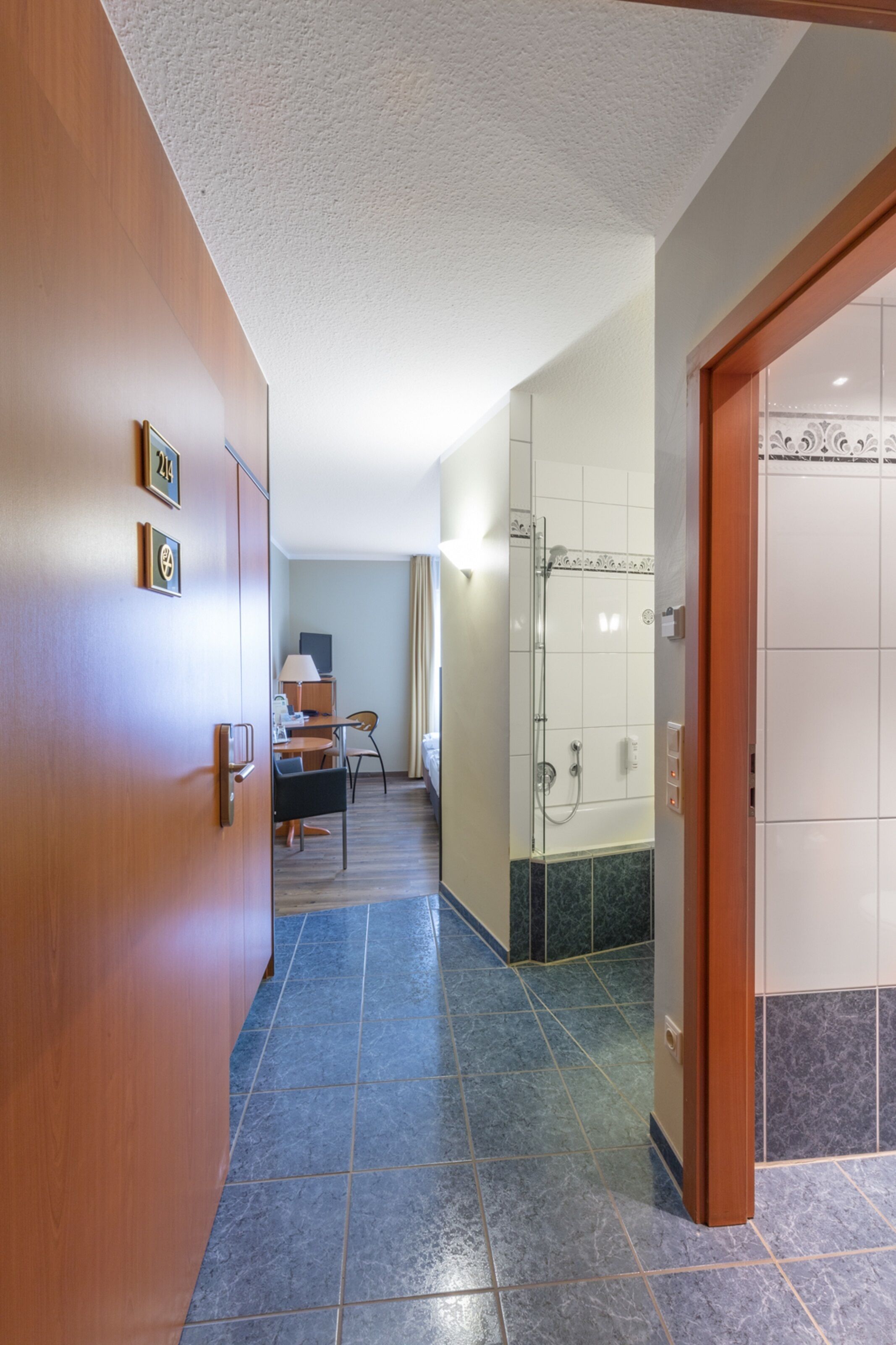 A hotel hallway with a wooden door, a desk with a chair, a shower area, and a tiled floor.