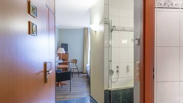 A hotel hallway with a wooden door, a desk with a chair, a shower area, and a tiled floor.