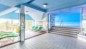 A sunlit indoor swimming pool area with green lounge chairs, a staircase leading to an upper deck, and a clear view of the sky through the ceiling. - Sport - und Tagungshotel De Poort (Goch)