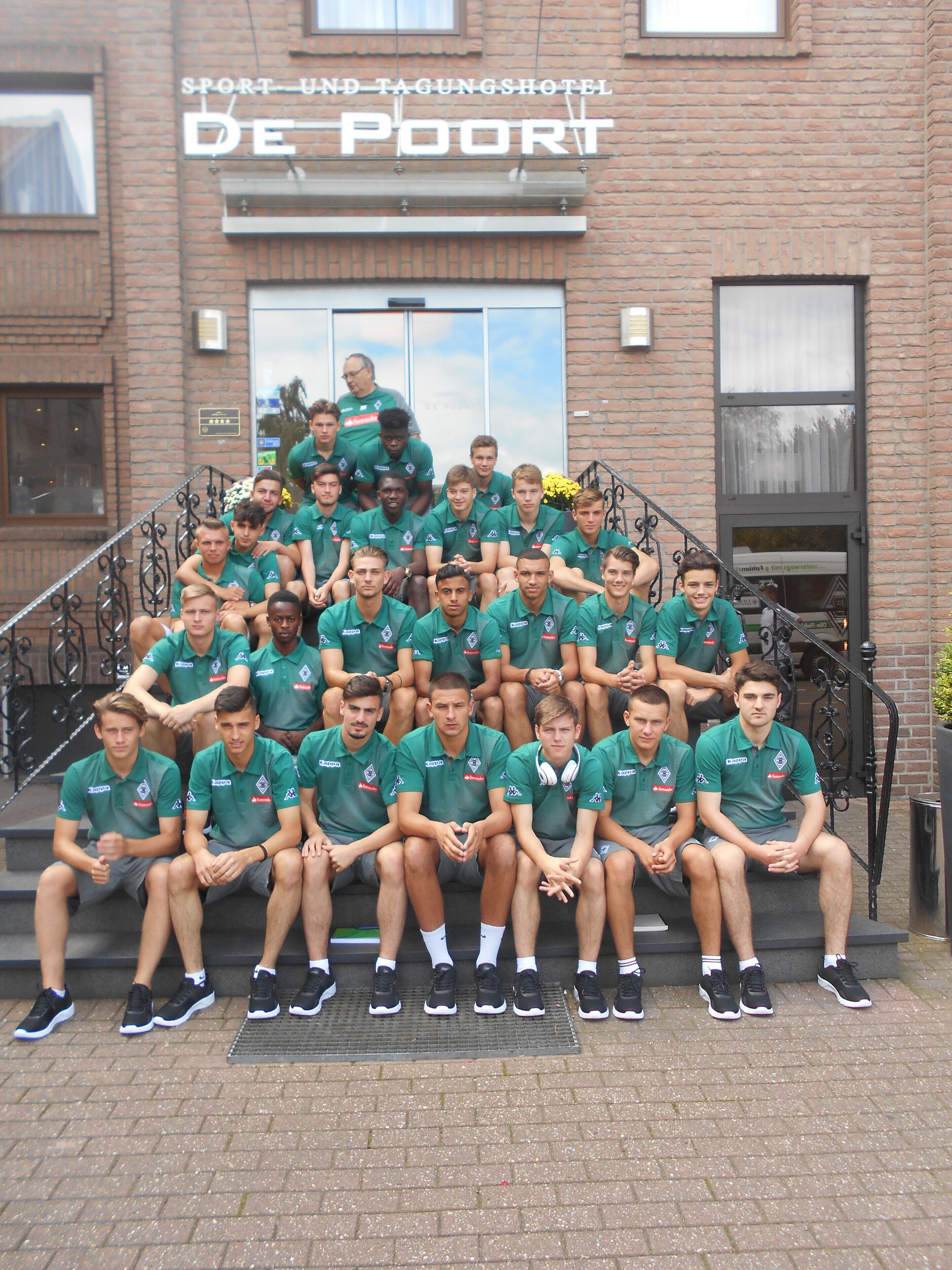 A group of people seated on steps in front of a building with a sign that reads "SPORT UND TAGUNGHOTEL DE POORT".