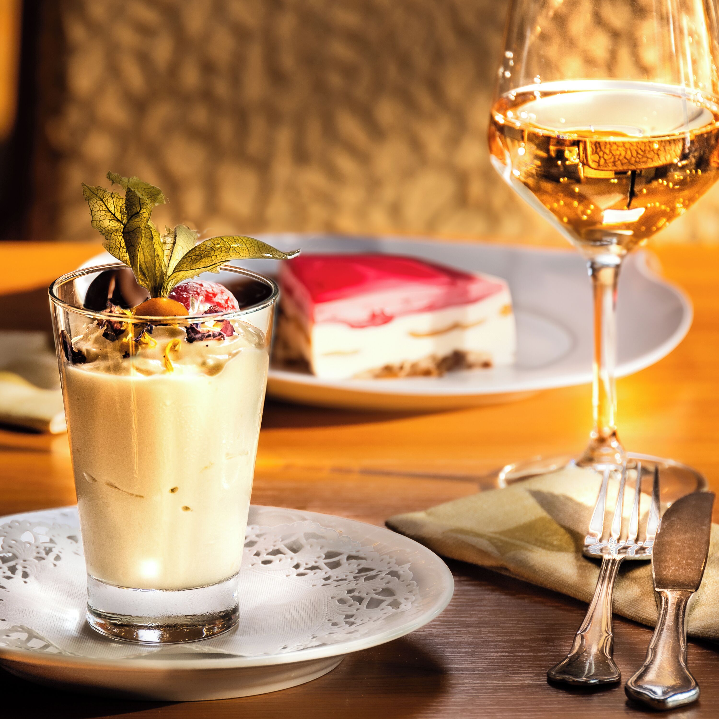 A dessert glass with a creamy dessert, a slice of cake on a plate, and a wine glass with a golden liquid.
