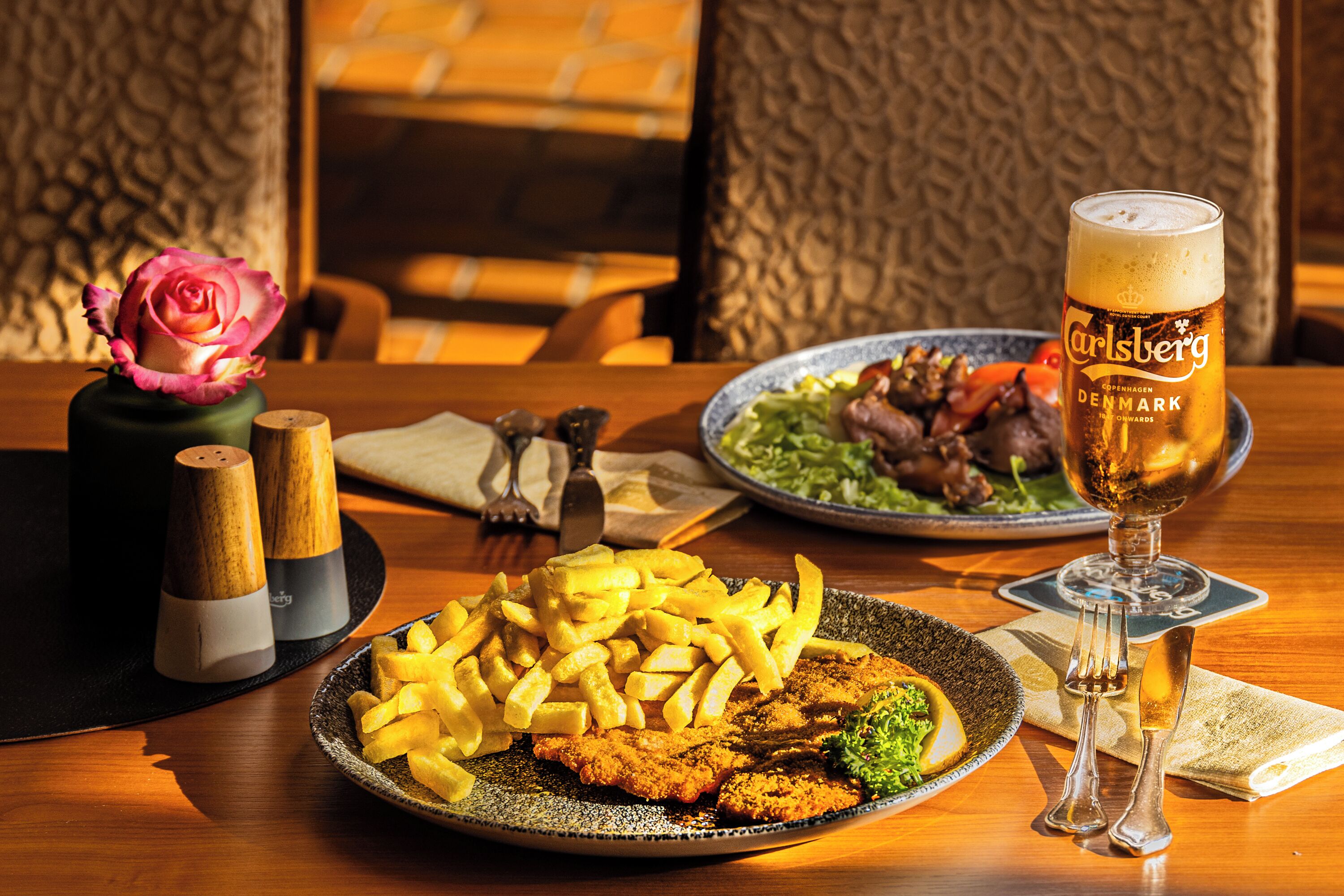 A dining table with a plate of food, a glass of beer, and a vase with a rose.