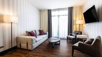 Ein Hotelzimmer mit einer Couch, Sesseln, einem Couchtisch, einem an der Wand montierten Fernseher und einem großen Fenster mit Vorhängen.