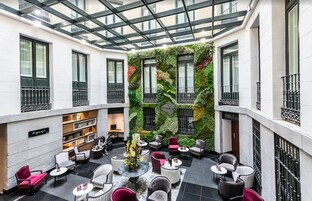 A modern hotel lobby with a glass ceiling, a green wall with plants, and a seating area with various chairs and tables.