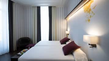 A hotel room with two beds, a chair, a small table with fruits, and a wall-mounted lamp.