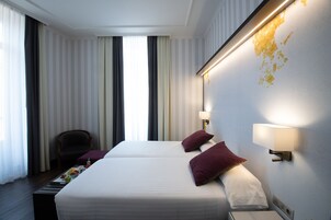 A hotel room with two beds, a chair, a small table with fruits, and a wall-mounted lamp.