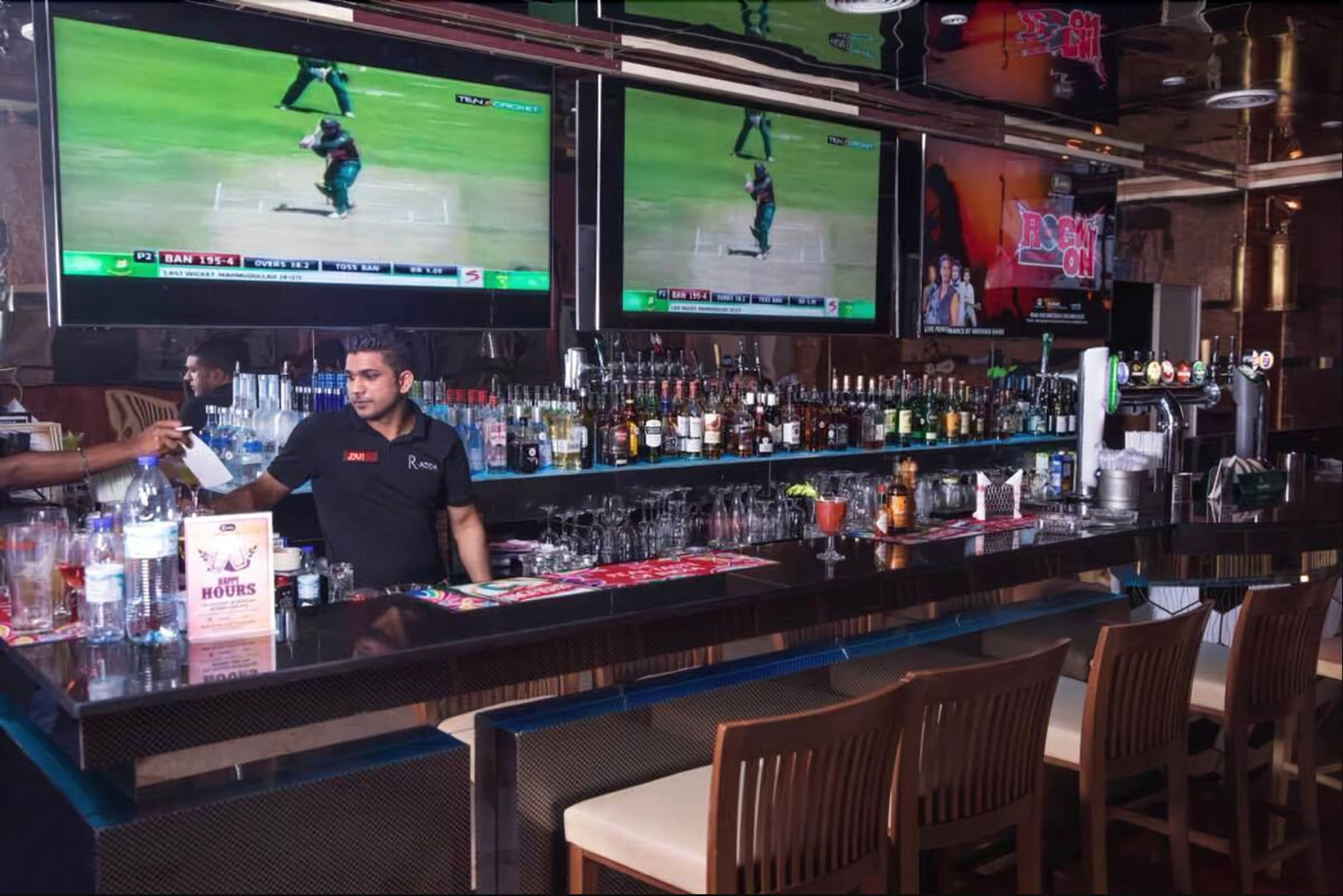A bar with multiple screens displaying cricket matches, a bartender, and a variety of bottles and glasses on the shelves.