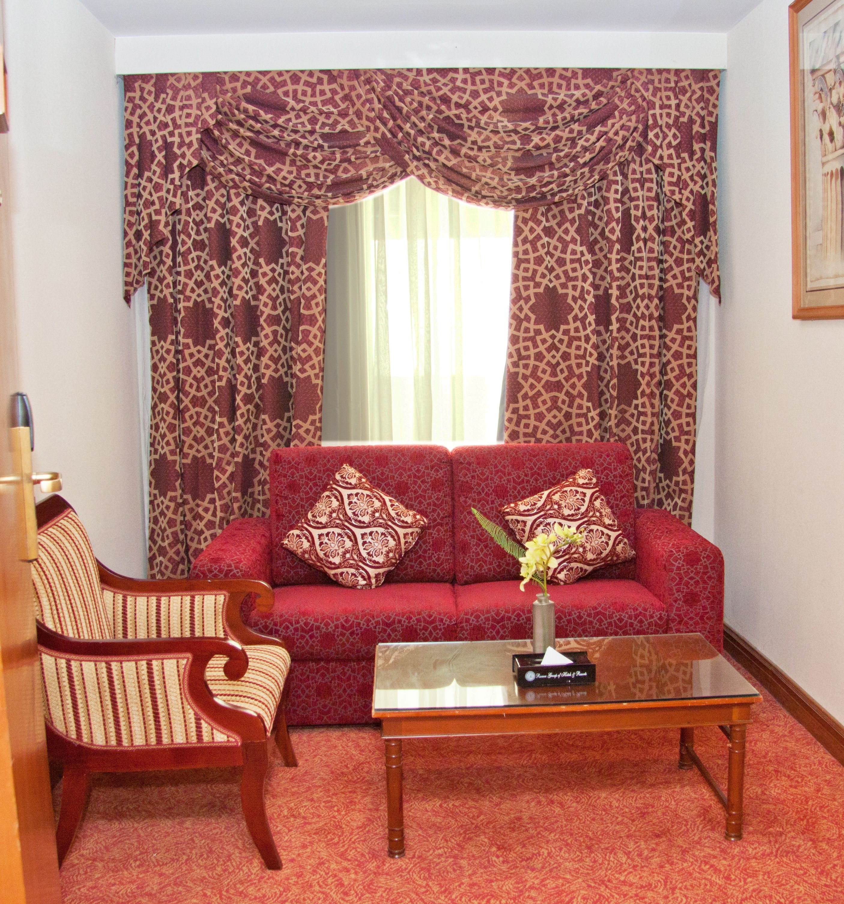 A living room with a red patterned sofa, two patterned cushions, a wooden chair, and a glass coffee table with a vase of flowers.