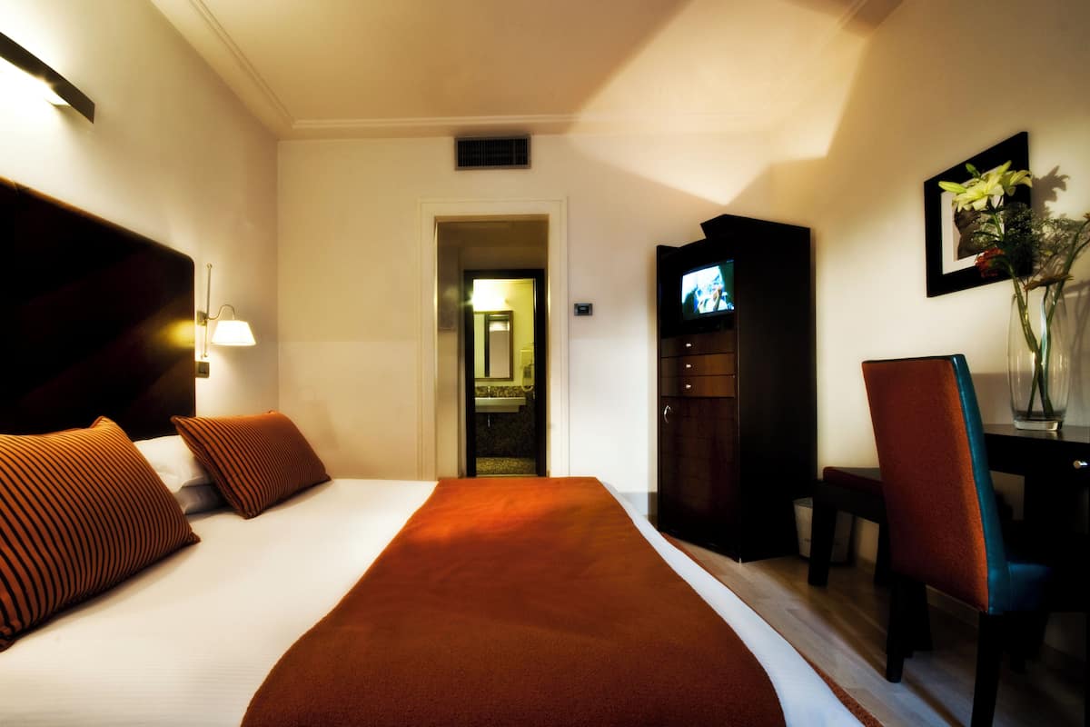 Ein Hotelzimmer mit einem großen Bett, einem Schreibtisch, einem Sessel, einem Fernseher und einer Blumenvase.