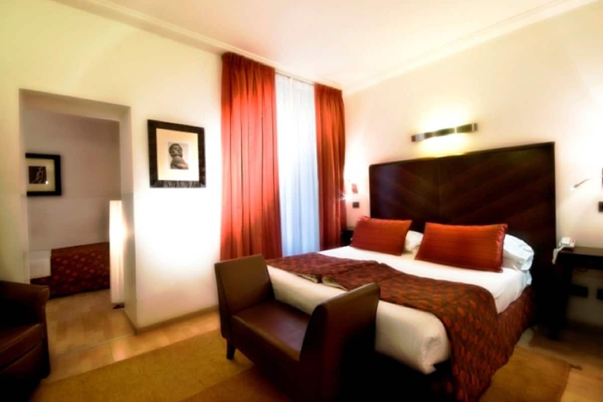 Ein Hotelzimmer mit einem großen Bett, zwei Kissen, einem Nachttisch, einem Sessel und einem gerahmten Bild an der Wand.